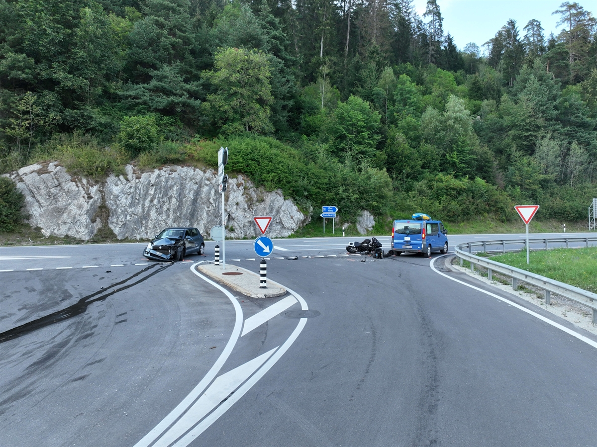 Strassenverlauf der Oberalpstrasse im Bereich der Unfallstelle in Schluein. Auf der linken Seite der beteiligte an der Front beschädigte dunkle Personenwagen. Rechts am Boden liegend das Motorrad, daneben ist der blaue Personenwagen der Folgekollision zu erkennen.