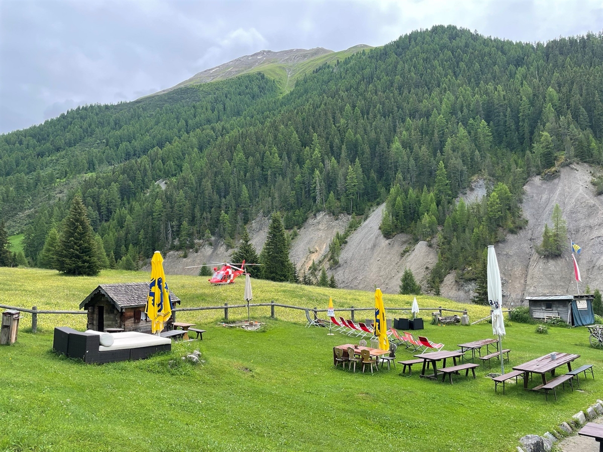 Der mit Bänken für Gäste zugängliche Bereich der Parkhütte Varusch. Auf der Wiese steht ein Rega-Helikopter. Blick in Richtung Berge und Nationalpark.