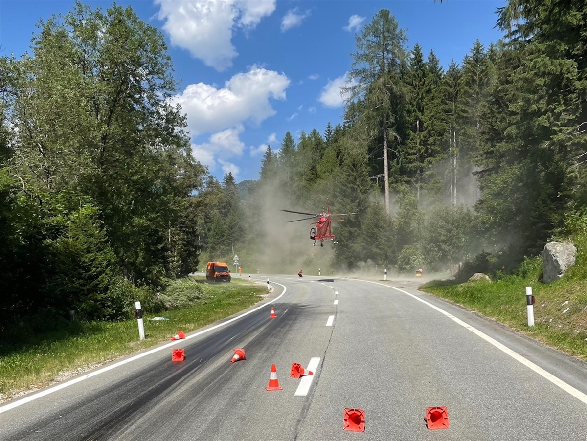 Die auf der einen Fahrbahnhälfte verschmutzte Strasse, umsäumt von Wald. Der Rega-Helikopter während der Landung.