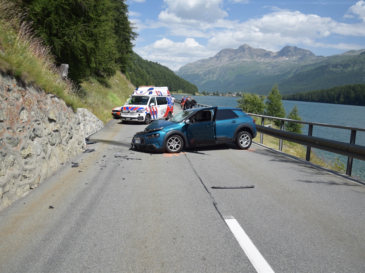 Das an der Front total beschädigte Auto steht quer auf der Strasse. Links die Stützmauer, rechts der Silsersee. Im Hintergrund ein Rettungswagen. Berge und grösstenteils heiterer Himmel.