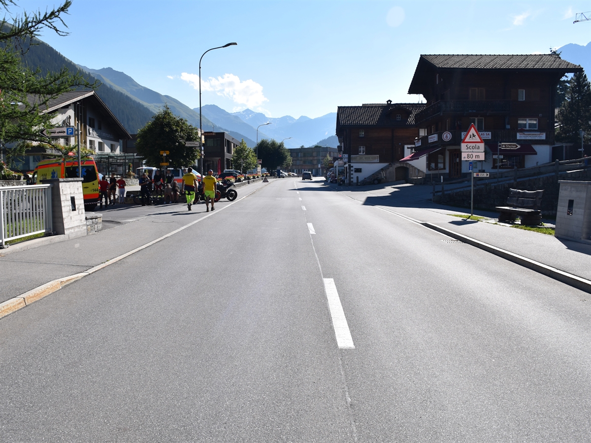 Die Oberalpstrasse in Richtung Pass gesehen. Am linken Bildrand ein Rettungswagen sowie viele Personen in unmittelbarer Nähe. Ebenfalls bei den Personen stehen Motorräder und am Boden liegt ein Fahrrad.