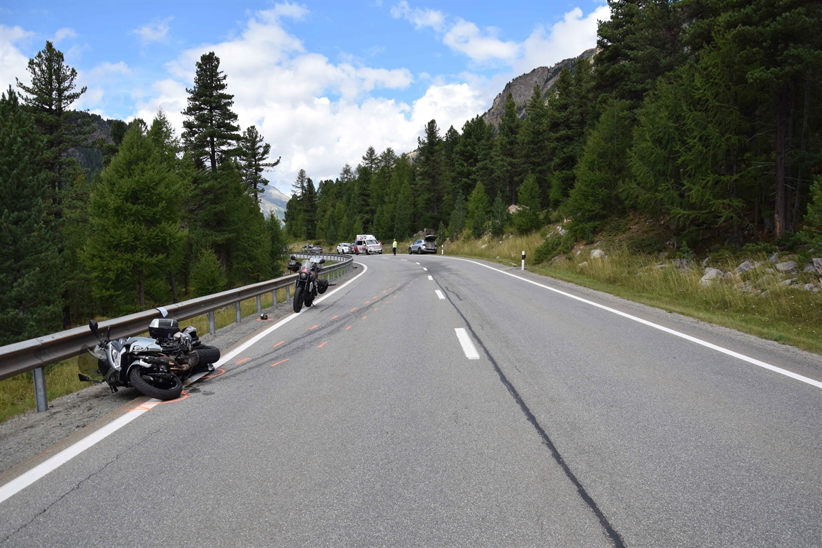 Übersicht der Unfallstelle. Linksseitig liegt das beteiligte Motorrad, dahinter steht ein nicht beteiligtes Motorrad auf dem Boden sind die roten Unfallmarkierungen zu sehen. Weiter hinten sind rechtsseitig am Strassenrand ein Ambulanzfahrzeug, das beteiligte Auto und weitere Fahrzeuge zu sehen.