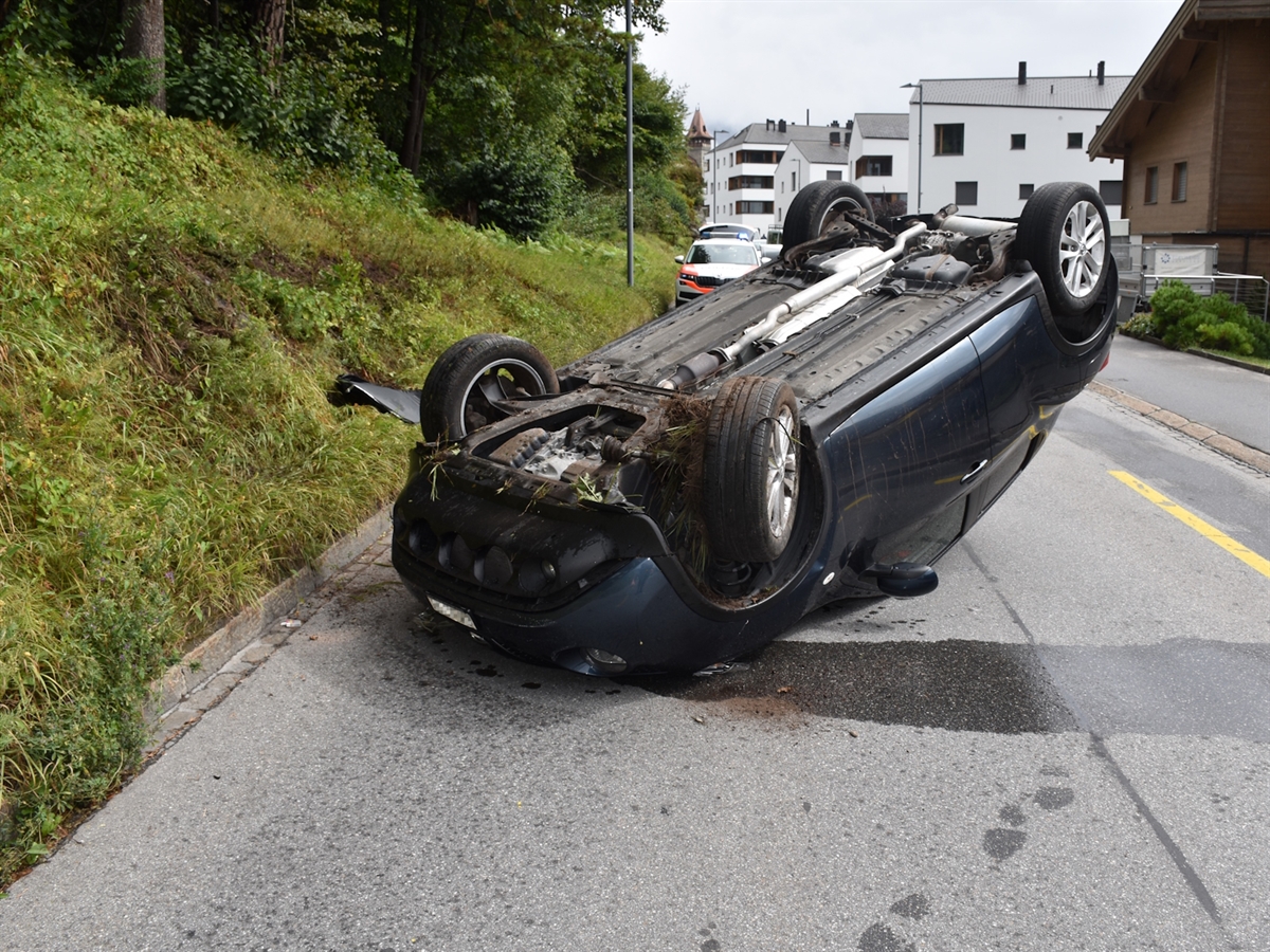 An der Böschung Kollisionsspuren des auf seinem Dach auf der Strasse liegende Autos. Im Hintergrund ein Polizeiauto und Häuser.