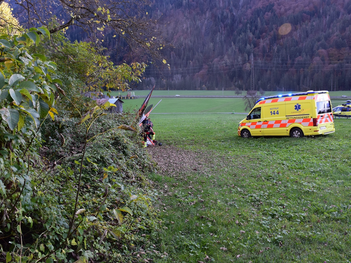 Hinter einem Heckenband liegt der abgestürzte Deltasegler auf der Wiese. Ebenfalls steht der Rettungswagen sowie der Helikopter der AAA Alpine Air Ambulance auf der Wiese. Rettungskräfte betreuen den Verletzten.