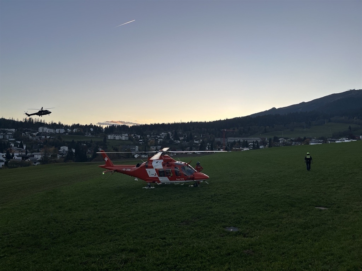 Beginnende Abenddämmerung. Ein Rega-Helikopter auf einer Wiese, dahinter in der Luft ein Armeehelikopter. Im Hintergrund das Dorf Flims und Berge.