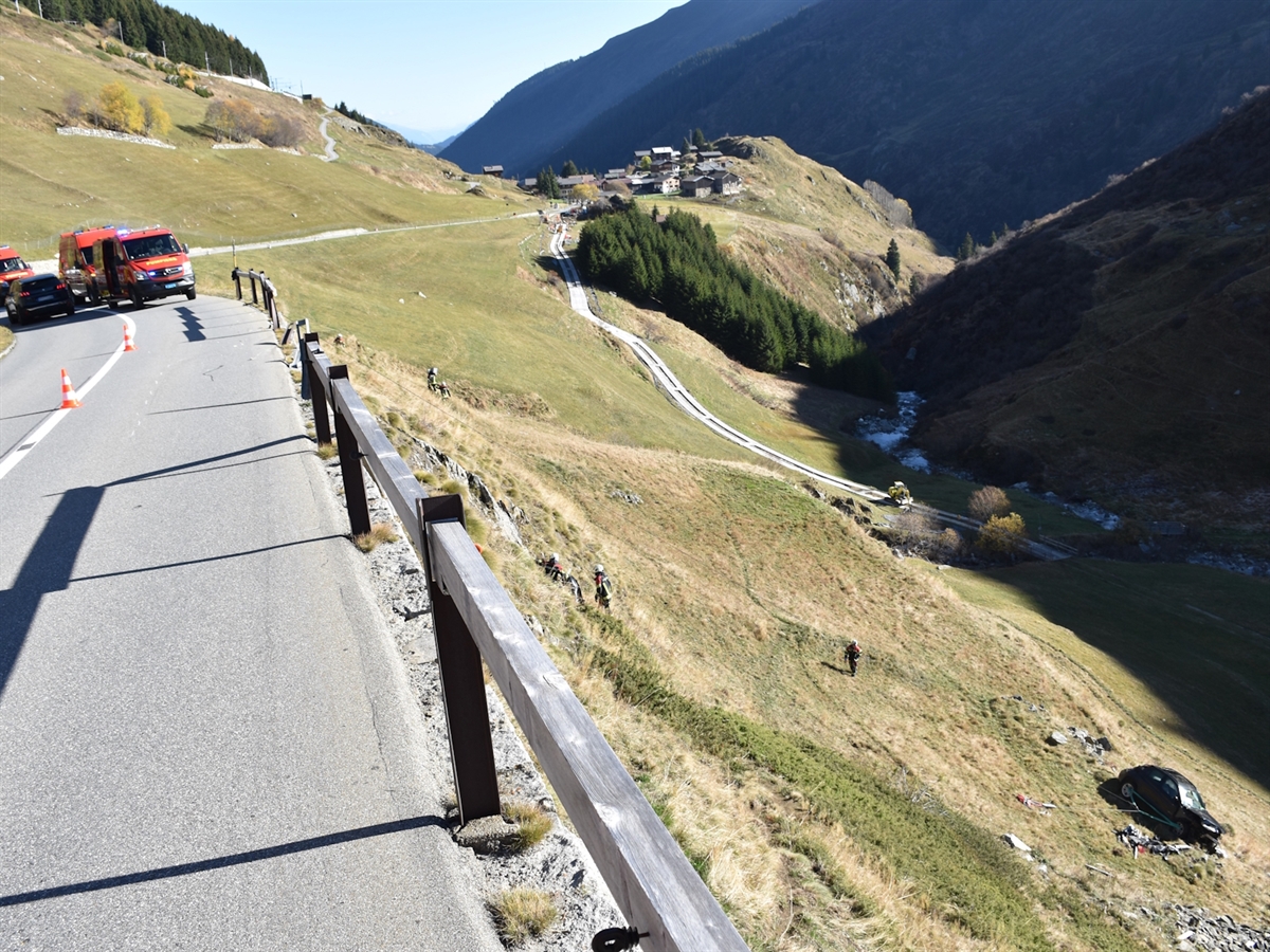 Die Oberalpstrasse mit durchbrochenem Bündnerzaun und Einsatzfahrzeugen der Feuerwehr. Rechts im abfallenden Hang das Auto und Feuerwehrleute.