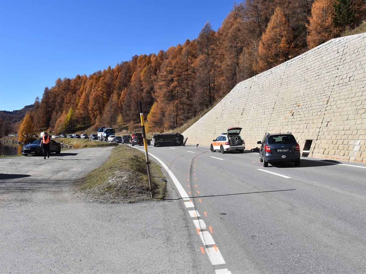 Rechts eines der Unfallautos nahe der Stützmauer. Dahinter ein Polizeiauto und das auf der Seite liegende Auto. Wolkenloser Himmel und herbstlich gefärbter Lärchenwald.