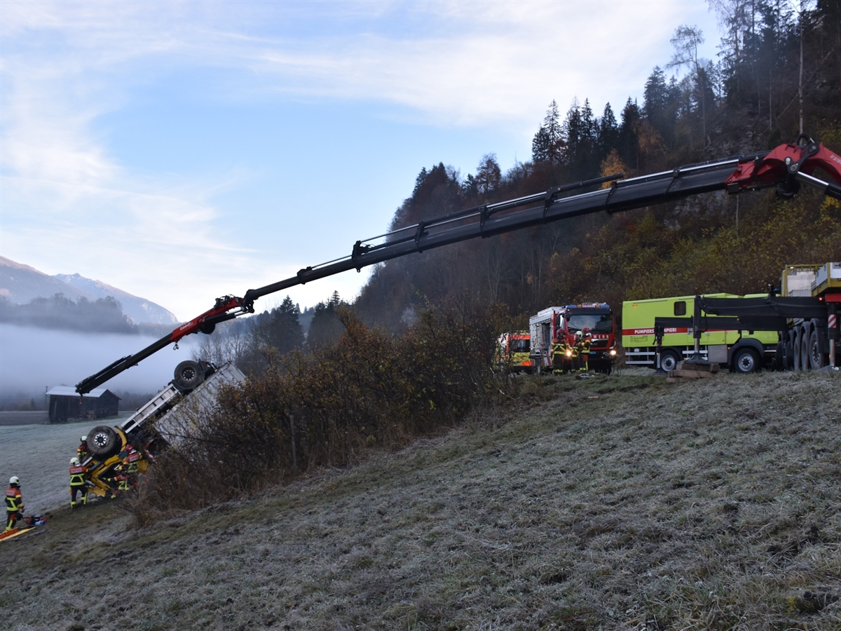 Links auf einer Wiese der auf dem Dach liegende Lastwagen. Er wird von einem Autokran gesichert. Im Bereich der Lastwagenkabine Einsatzkräfte während der Bergungsarbeiten.