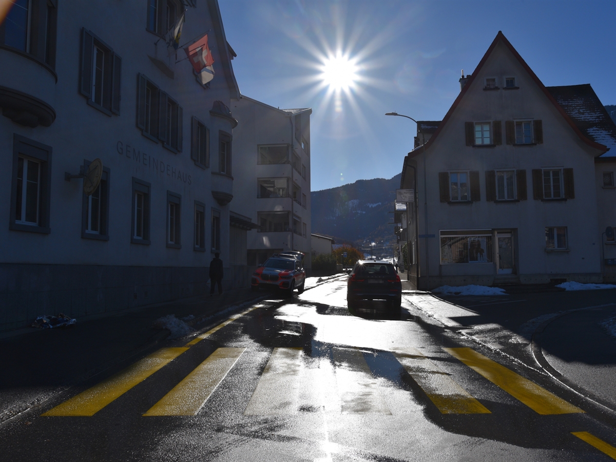 Die Italienische Strasse wird durch die tiefstehende Sonne bestrahlt. Die nasse Fahrbahn spiegelt die Sonne. Auf der Fahrbahn das Unfallauto. Hinter diesem der Fussgängerstreifen.