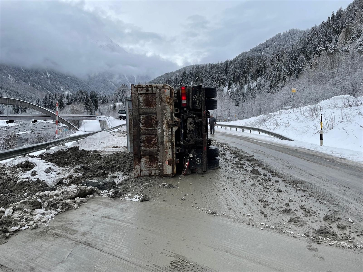 Der auf der linken Seite mitten auf der Strasse liegende Lastwagen von hinten. Die Strasse ist mit dem verschütteten Aushubmaterial verunreinigt.