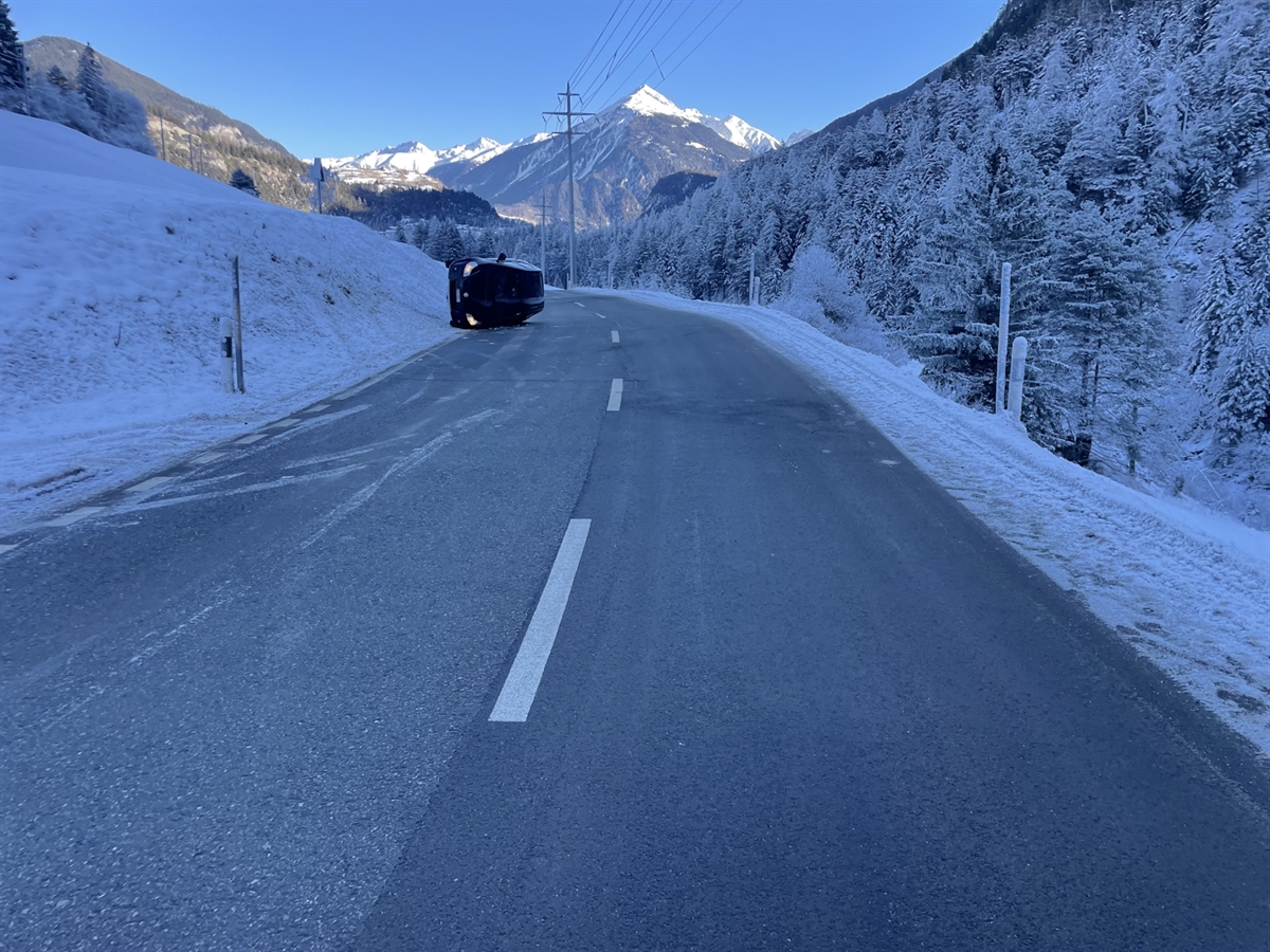 Das Unfallauto liegt auf der linken Fahrzeugseite am linken Strassenrand. Das Bild der Winterlandschaft wird mit besonnten Bergen und blauem Himmel abgerundet.