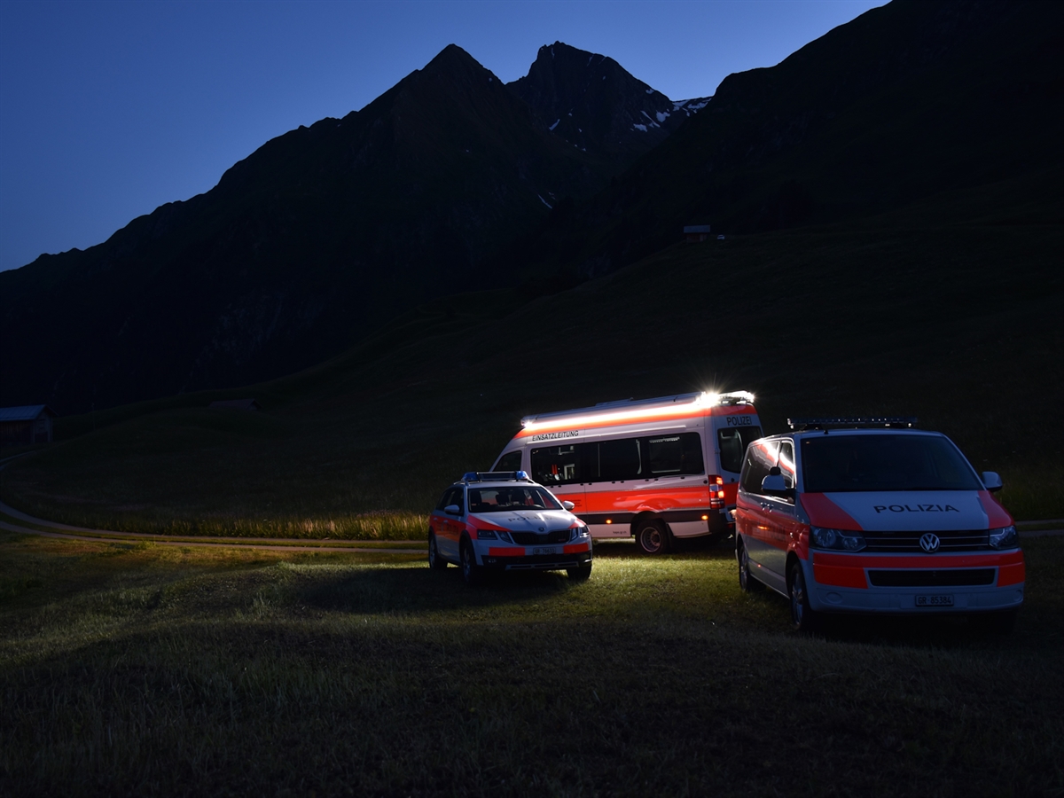 Das beleuchtete Einsatzleitfahrzeug der Kapo GR mit zwei Patrouillenfahrzeugen stehen in der Abenddämmerung vor einer Bergkulisse, welche mit dunkelblauem Himmel umrahmt wird.