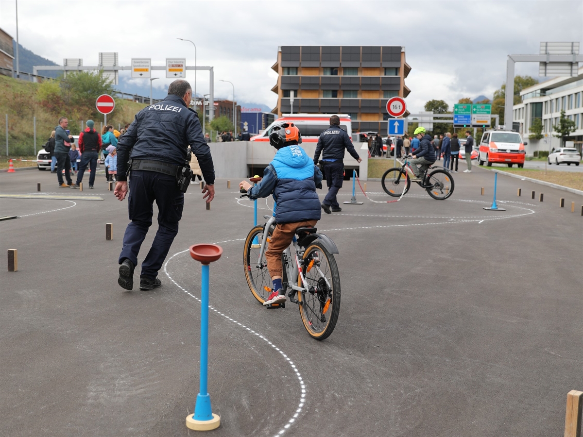 Ein Kind umkurvt mit dem Velo diverse Hindernisse. Im Hintergrund Publikum, das Gebäude und Polizeiautos.